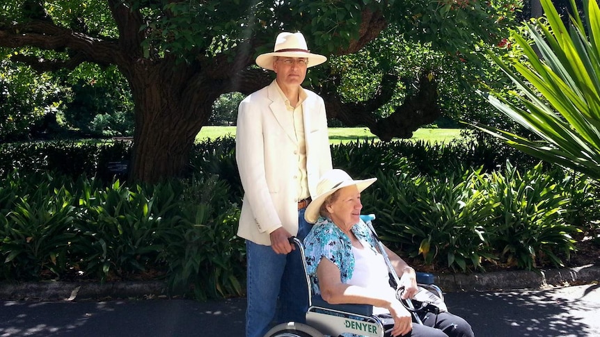 A man in a linen jacket and panama hat stands behind a woman in a hat, sitting in a wheelchair, surrounded by a lush garden.