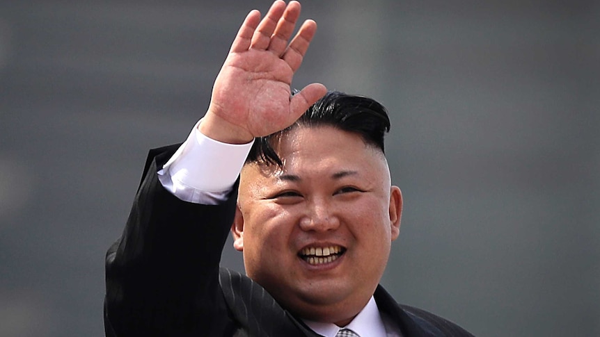 A midshot of North Korean leader Kim Jong Un, wearing a suit. He is waving and smiling.