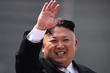 A midshot of North Korean leader Kim Jong Un, wearing a suit. He is waving and smiling.