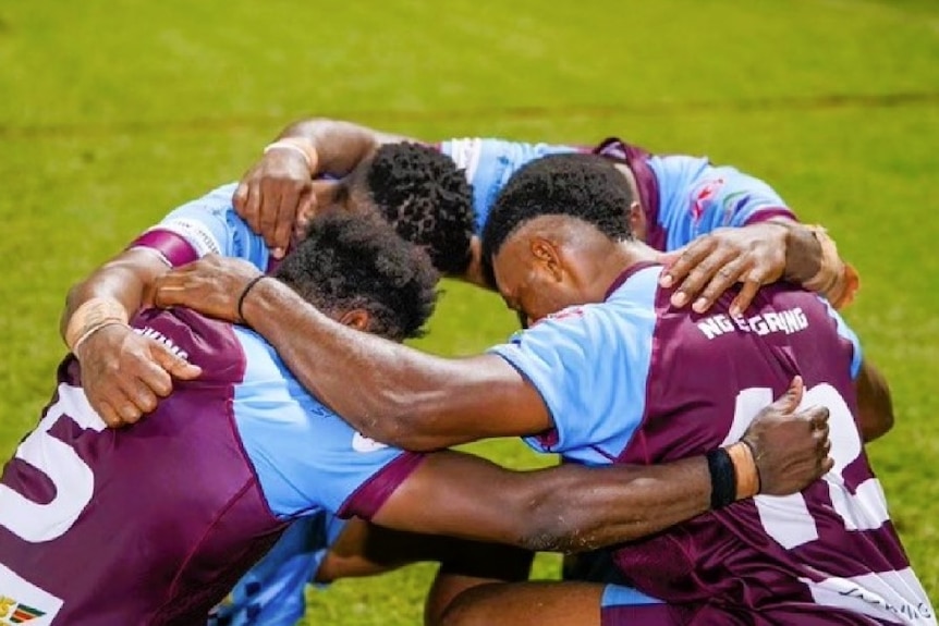 Four men from Papua New Guinea wearing maroon and sky blue football jerseys kneeling on a field and praying.