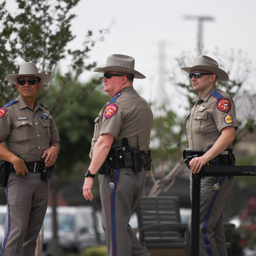 Three US police officers standing earing beige-grey uniforms with cowboy hats.