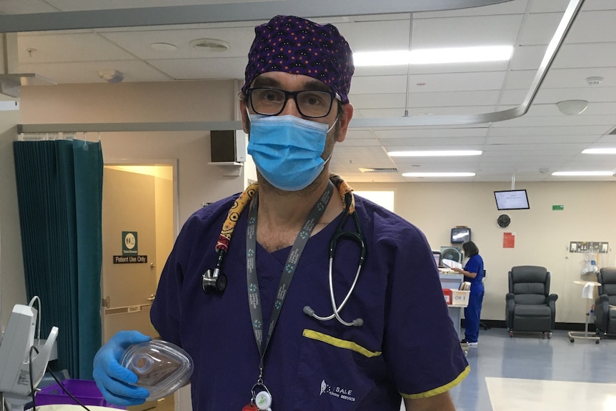 A man stands in a hospital corridor wearing doctor's scrubs, a stethoscope, glasses and a face mask.