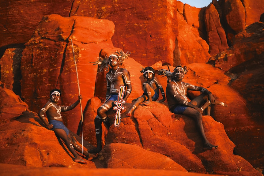 Sitting on bright red rocks, two Indigenous children and men wear traditional body paint and head-dress, looking into distance.