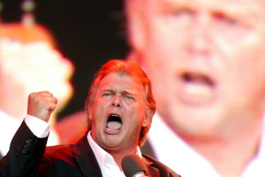 John Farnham singing on stage in a black suit with a large video screen behind him on the stage.