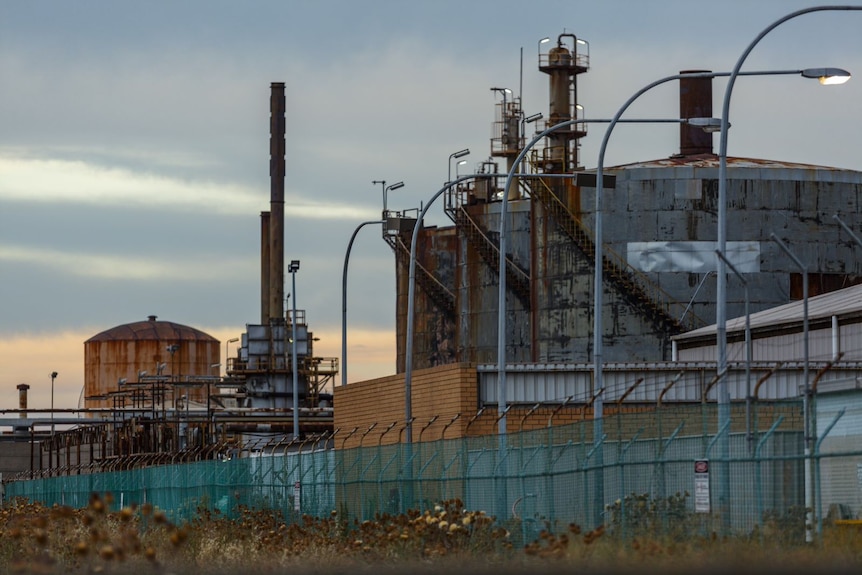 Three big rusted tanks and a chimney can be seen in the fading light