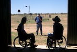 two silhouetted farmers in wheelchair in the door of a shed look out on another man standing near a windmill.