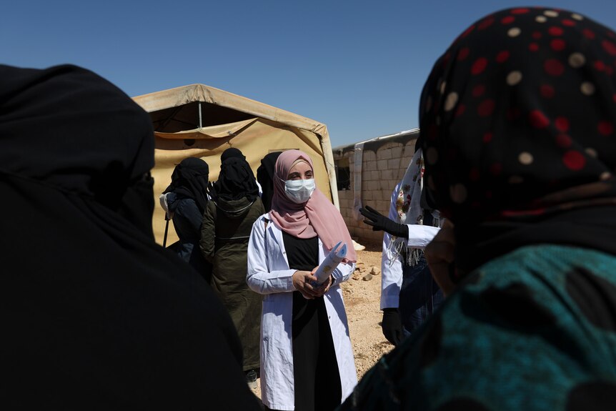 A doctor wearing a facemask and headscarf stands in a group of women and children. 