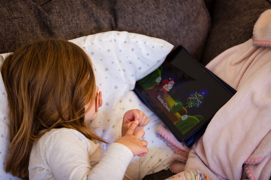 Three-year-old Harlow lies on the couch watching a show on an ipad. 