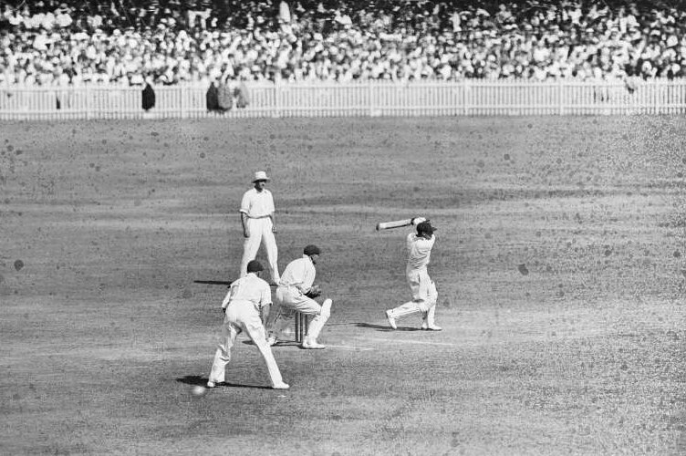 A man faces a delivery in a cricket game, surrounded by fielders