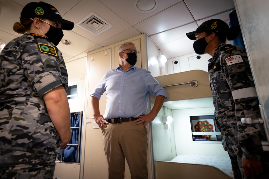 Scott Morrison wearing a face mask with his hands on his hips standing between two uniformed Navy personnel on a patrol boat