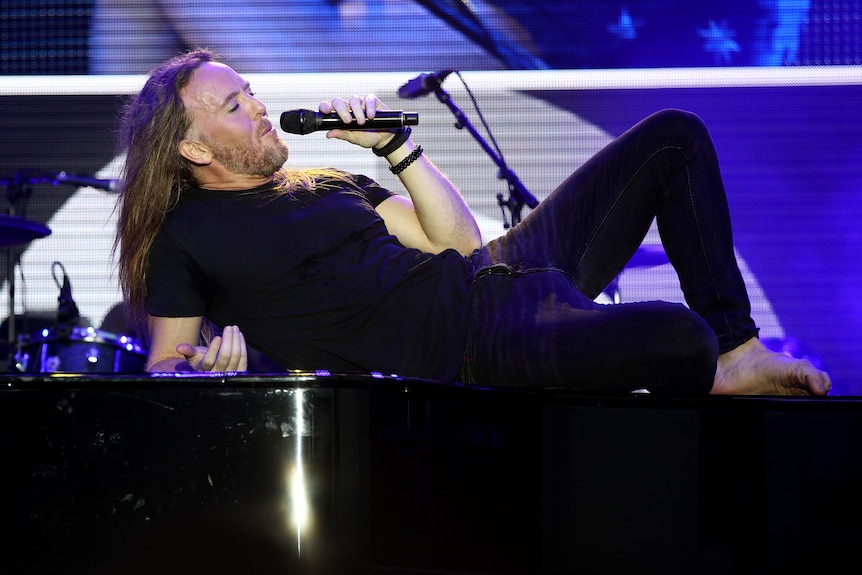 A man sings into a microphone while laying on top of a piano.