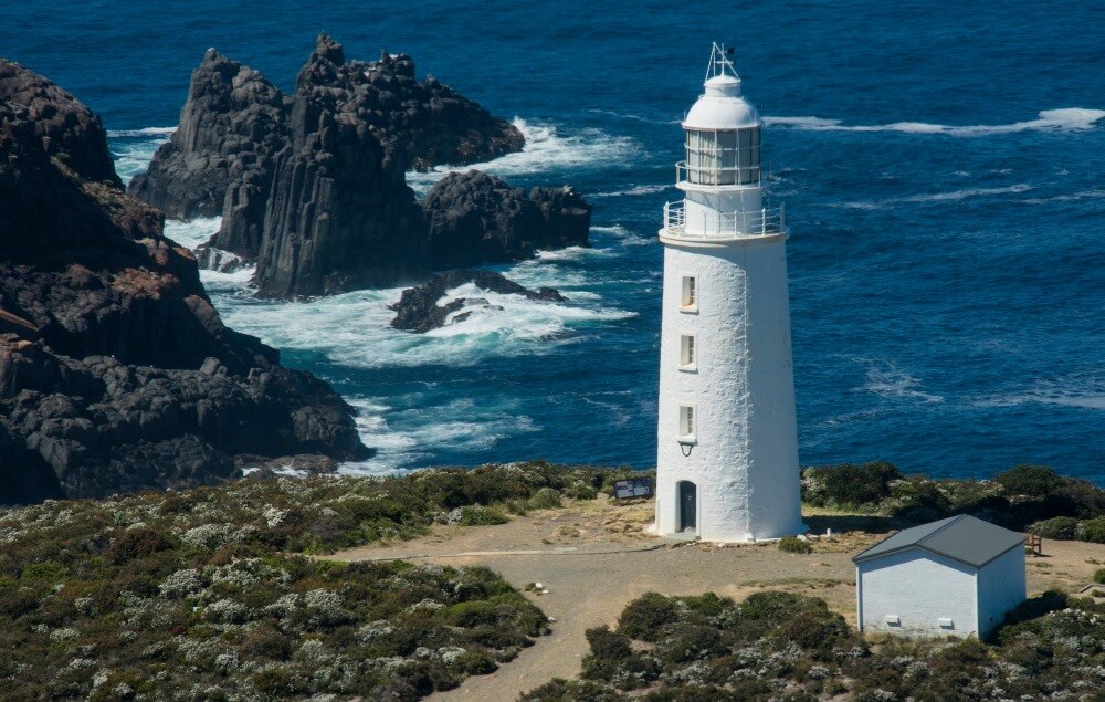 Colour photo of a white lighthouse with rocks and big swells