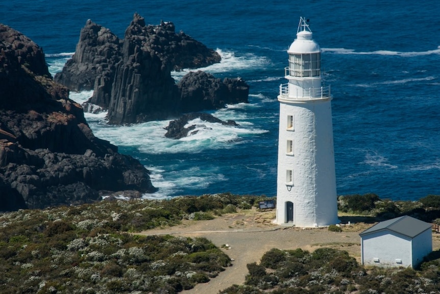 Colour photo of a white lighthouse with rocks and big swells