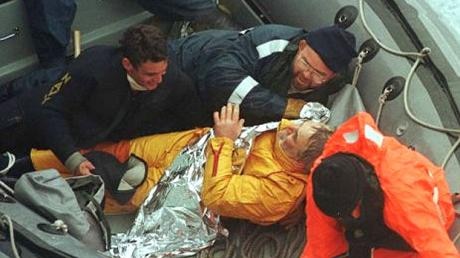 Four people in an inflatable boat, one wrapped in a insulating blanket.