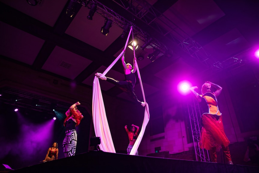 A stage of performers, including one hanging from silks.