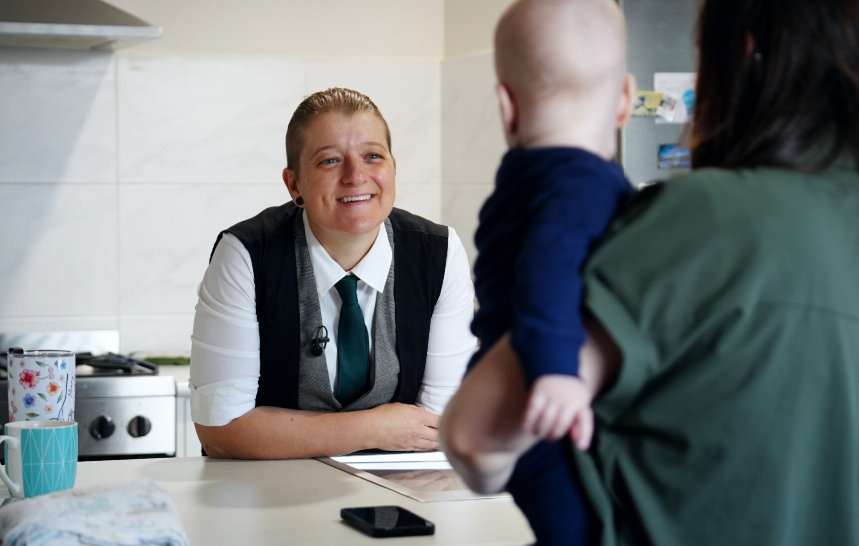 Una mujer vestida con camisa blanca, corbata negra y chaleco se apoya en un banco de la cocina y habla con una mujer que sostiene a un niño de espaldas.
