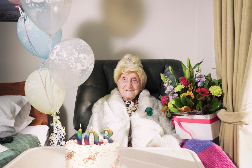 An elderly woman wears a fur coat and tiara in front of a birthday cake and balloons. 