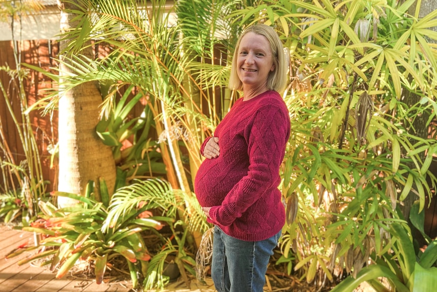 A woman stands in a garden, holding her pregnant belly. 