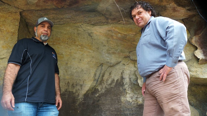 Two men standing in front of rock art with cracking down the side.