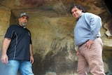 Two men standing in front of rock art with cracking down the side.