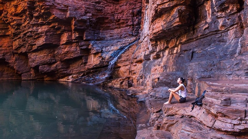 A woman sits by the water at the bottom of a gorge.