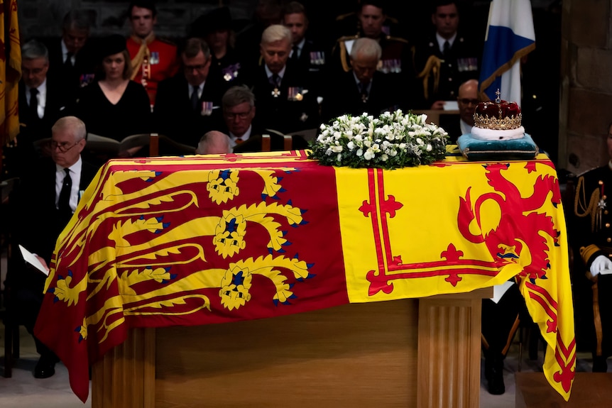 A crown sits on a coffin draped in a red and yellow flag.