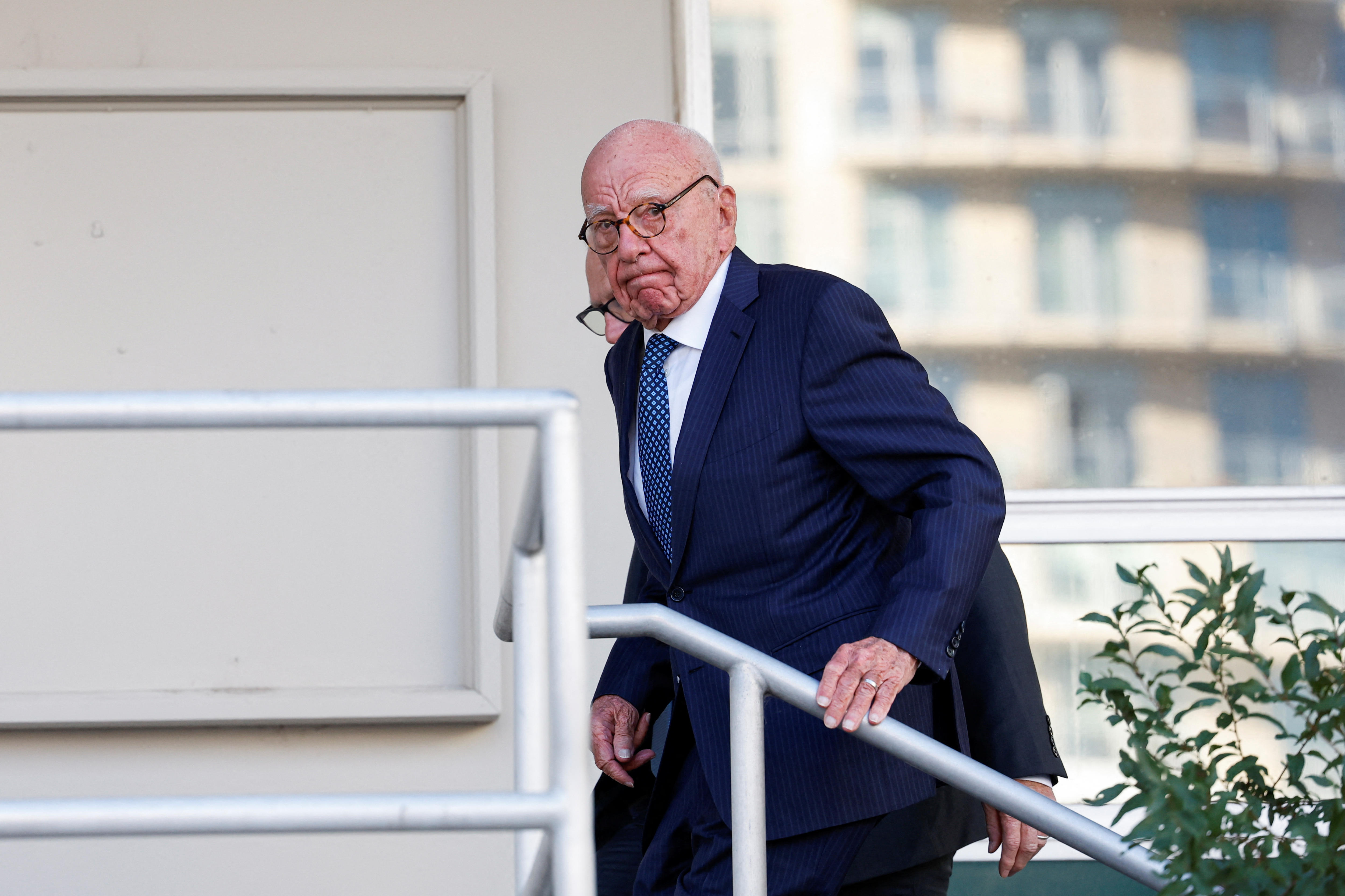 Rupert Murdoch walks up the stairs while holding onto the handrail with one hand.