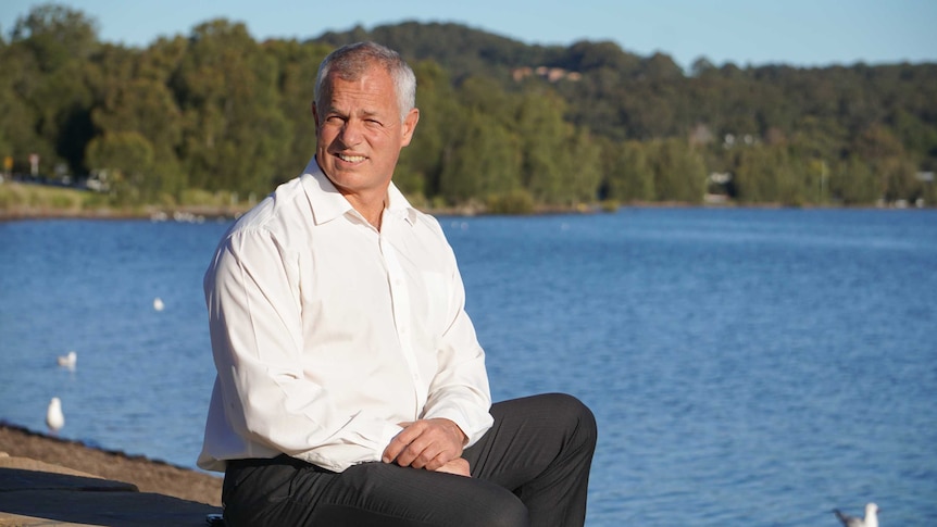 A man in a white shirt sitting in front of the water and looking at the camera