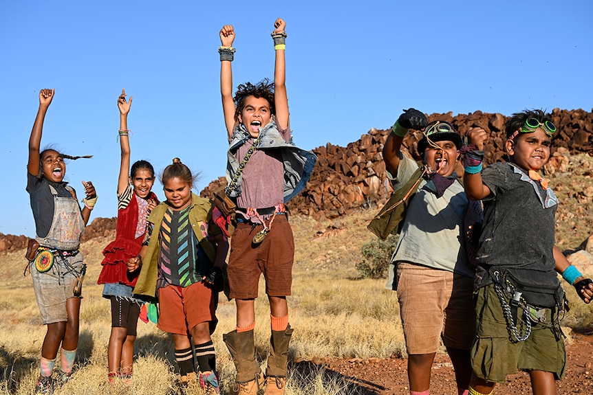 Six children raise hands and fist in the air and celebrate, one boy is jumping, in rocky desert landscape with spinifex.