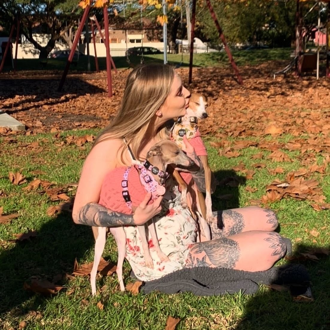 A woman sits on the ground, holding two Italian greyhound dogs.