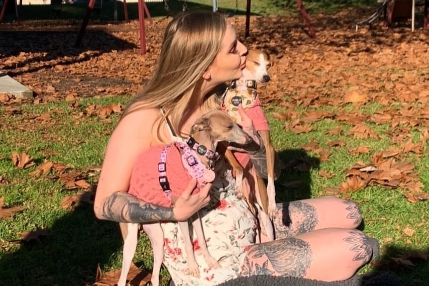 A woman sits on the ground, holding two Italian greyhound dogs.