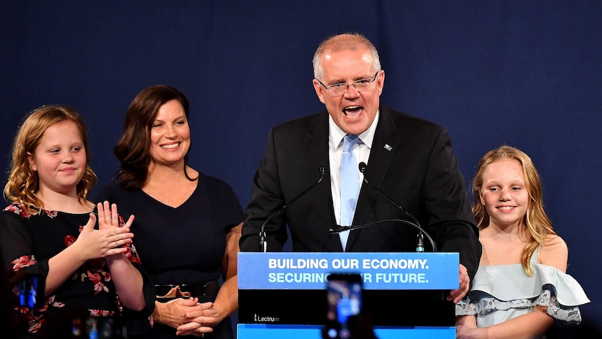 Scott Morrison talks at a podium surrounded by his smiling wife and daughters