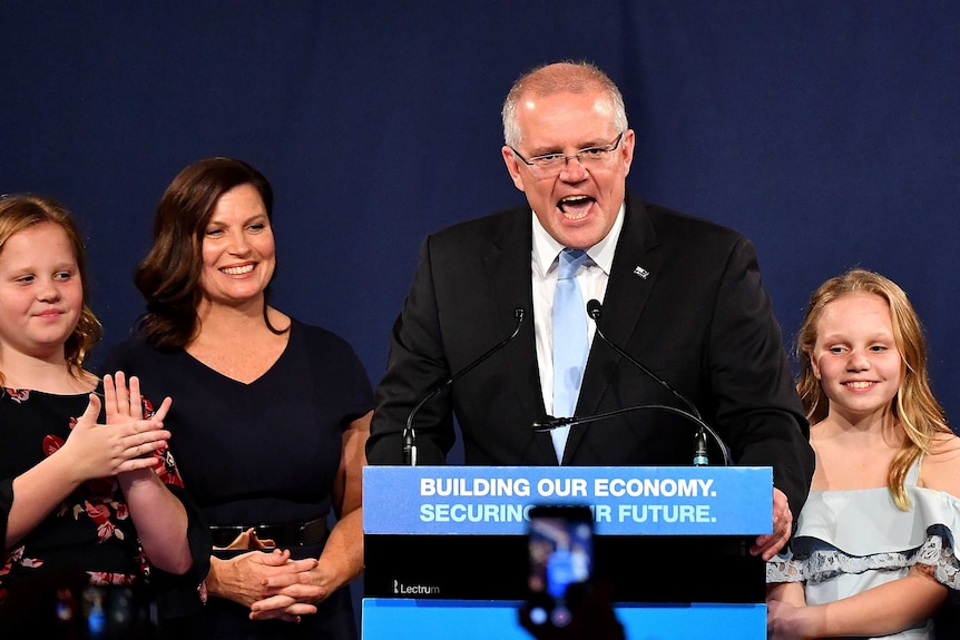 Scott Morrison talks at a podium surrounded by his smiling wife and daughters