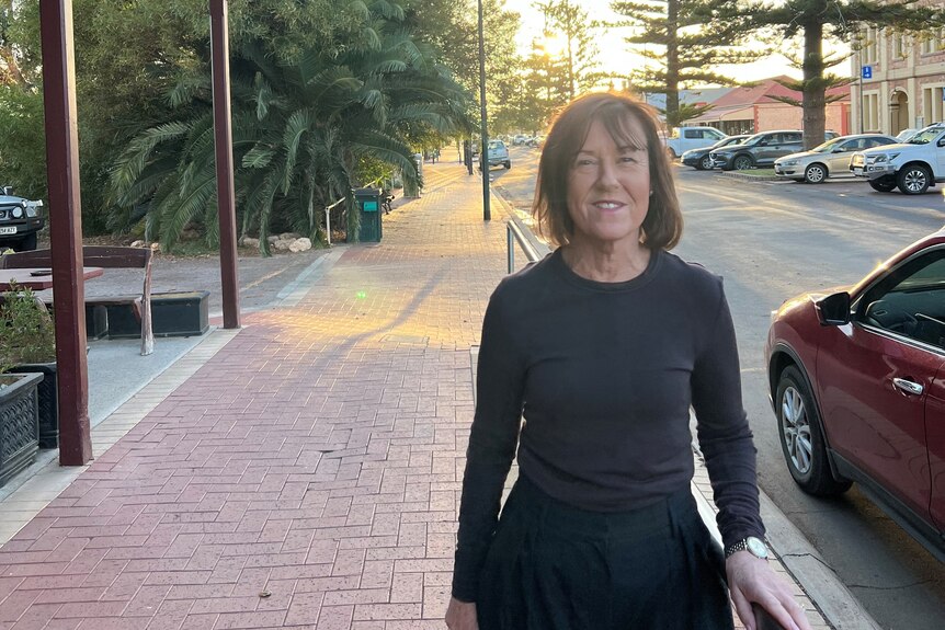 A woman in a black shirt stands in front of a street with a red car in background. 