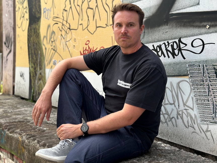 A man sits on an outdoor wall looking serious.