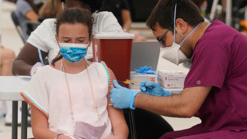 A teenage girl wearing a mask sits in a chair as a male administers her vaccination.