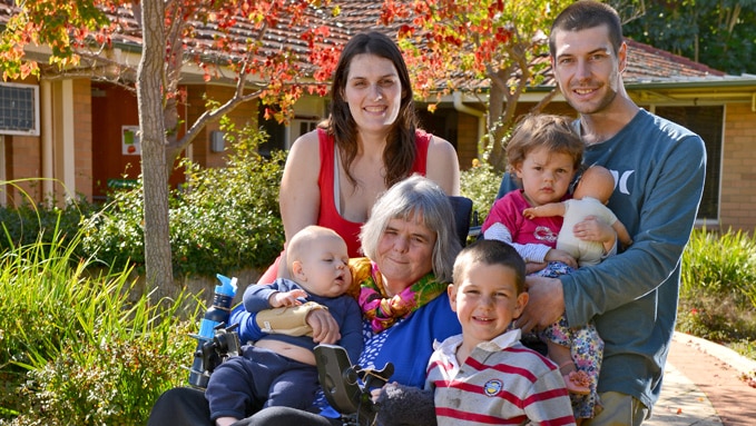An older woman in a wheelchair surrounded by her children and grandchildren, with a house and trees in background.