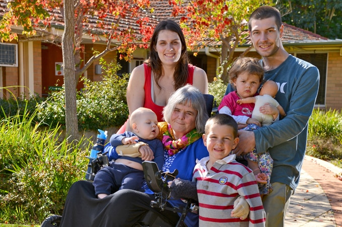 An older woman in a wheelchair surrounded by her children and grandchildren, with a house and trees in background.