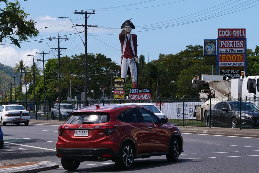 A large statue of a man in red coat and hat, stands with his arm extended, cars drive along the highway in front