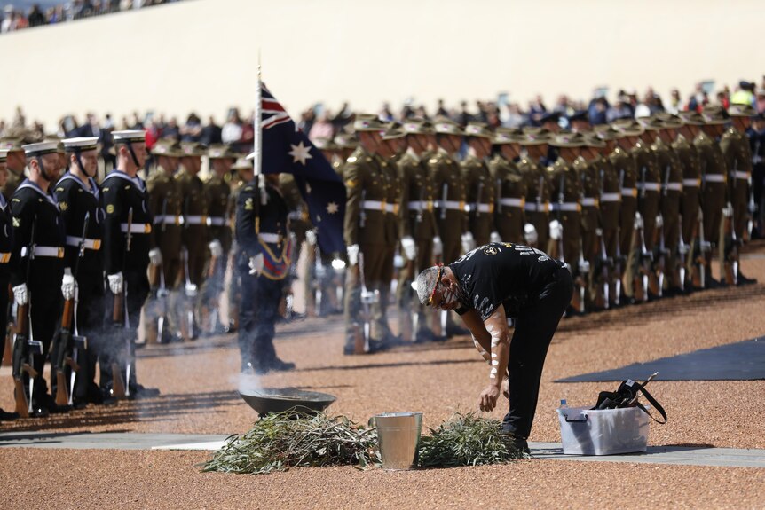 A man in traditional body paint lays branches in a smoking dish before rows of military personnel at Parliament House.