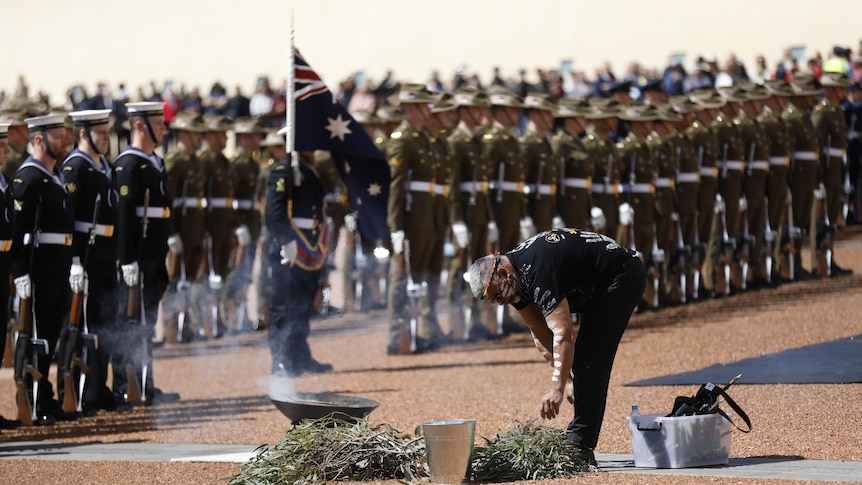 A man in traditional body paint lays branches in a smoking dish before rows of military personnel at Parliament House.