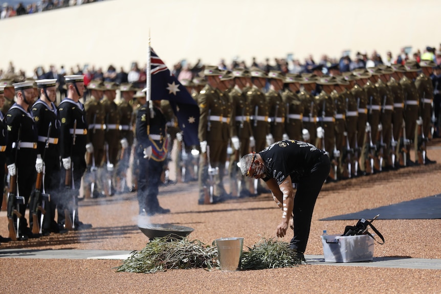 A man in traditional body paint lays branches in a smoking dish before rows of military personnel at Parliament House.