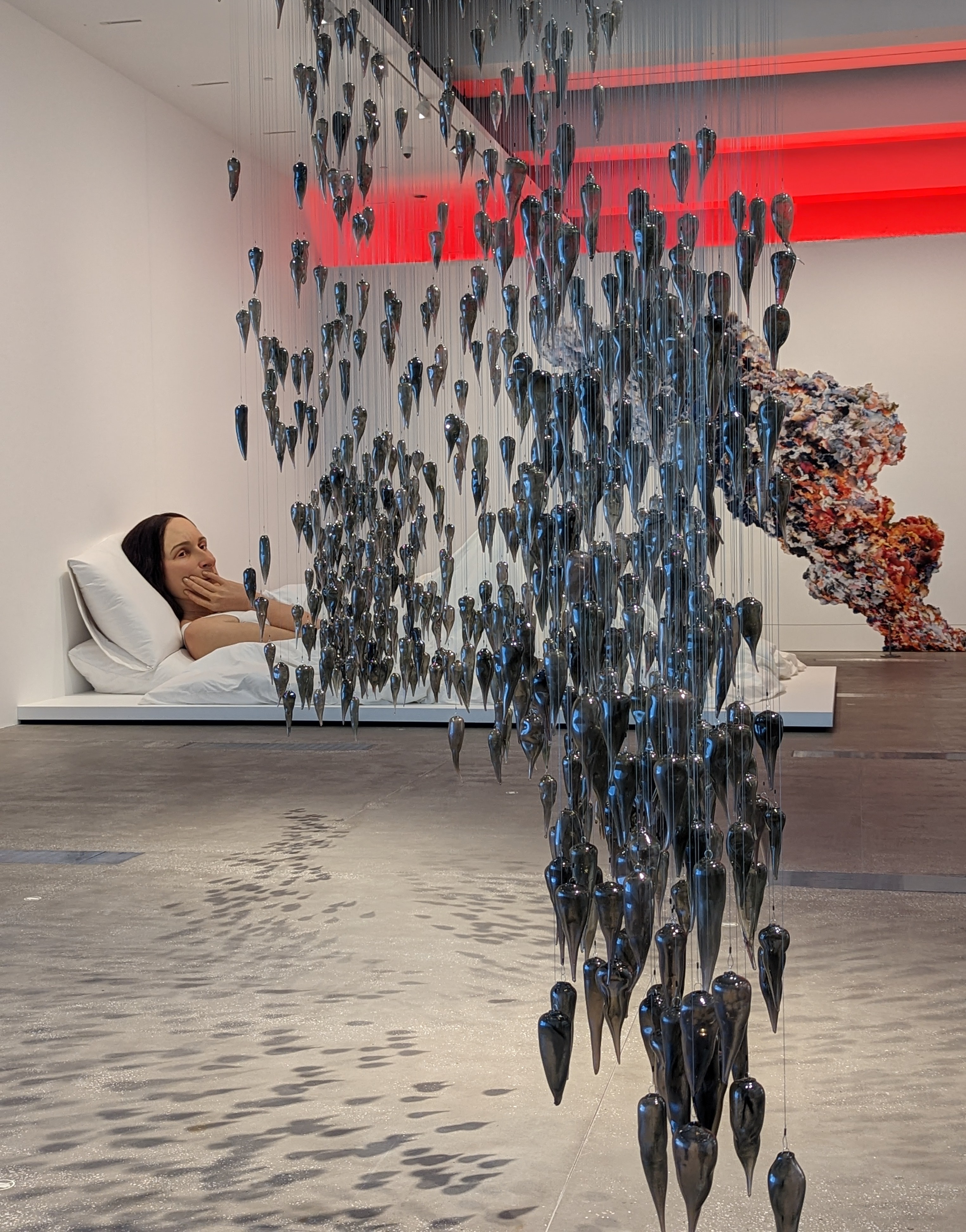 Glass yams hang from a gallery ceiling. Behind it is a large hyperrealistic sculpture of a woman, and a collaged plume of smoke.