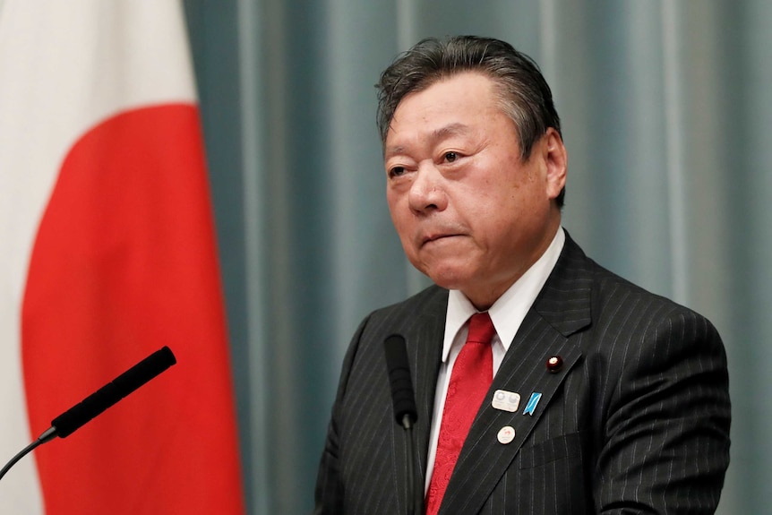 Japanese minister Yoshitaka Sakurada, wearing a suit, stands at a microphone to give a speech