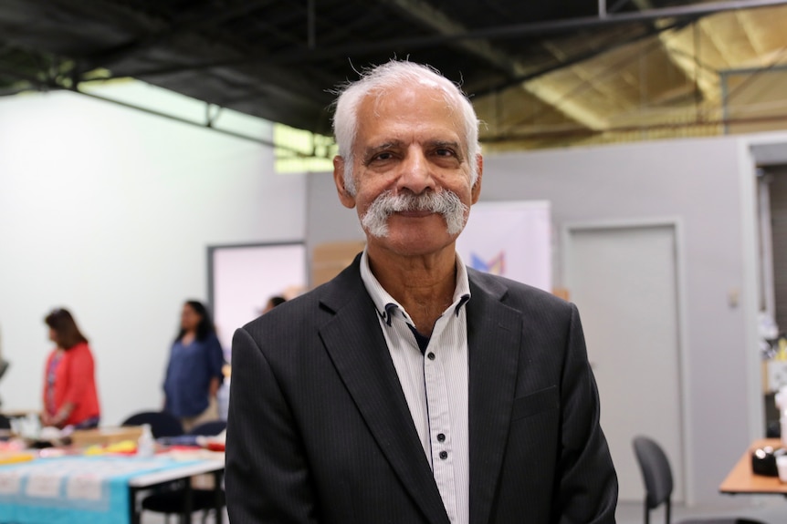 A mid-shot of a smiling man indoors with grey hair and a thick grey moustache, wearing a dark suit and white shirt.