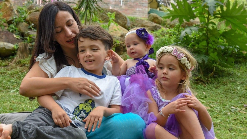 A dark-haired young mother hugs her son as the pair sit with a baby and a girl of a bout four in a garden.