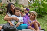 A dark-haired young mother hugs her son as the pair sit with a baby and a girl of a bout four in a garden.