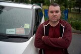 Michael Bradshaw, an Uber and DiDi driver in Ipswich, beside his car showing ride share app stickers and his arms folded
