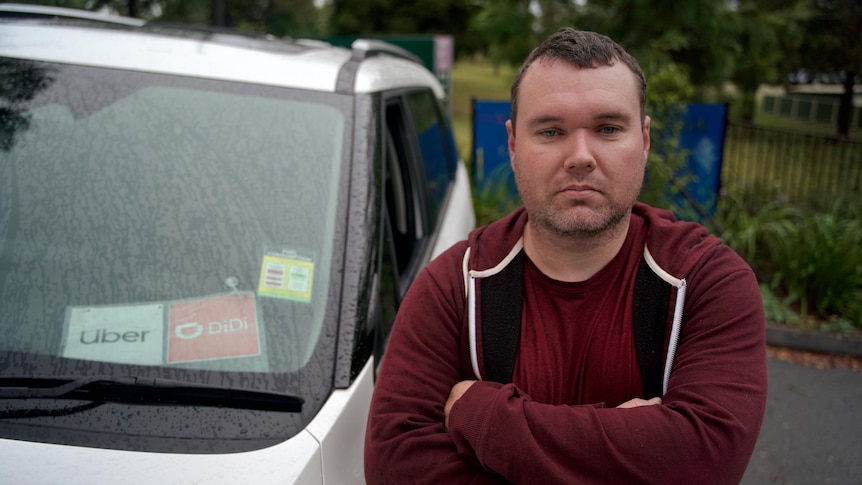 Michael Bradshaw, an Uber and DiDi driver in Ipswich, beside his car showing ride share app stickers and his arms folded