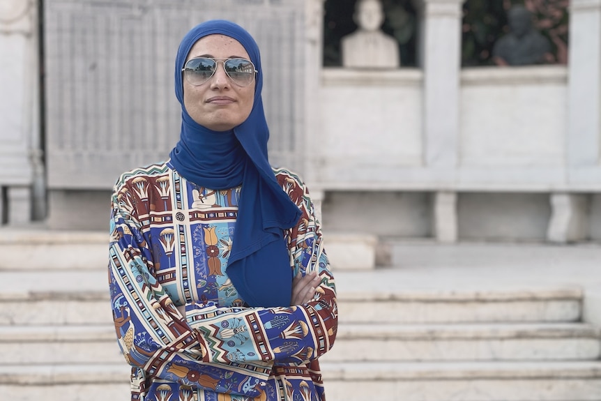 An Egyptian woman in a hijab and sunglasses, with a colourful top that has ancient Egyptian symbols.
